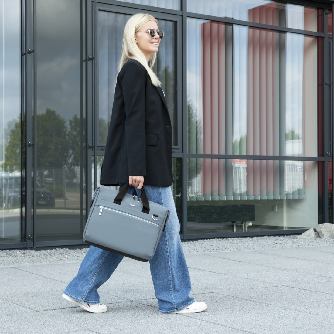 Frau geht, hält graue Tasche, trägt Blazer und Jeans. Glasgebäude im Hintergrund.