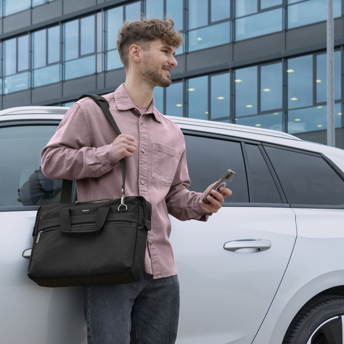 Ein Mann in einem rosa Hemd hält ein Telefon und eine schwarze Aktentasche neben einem Auto.