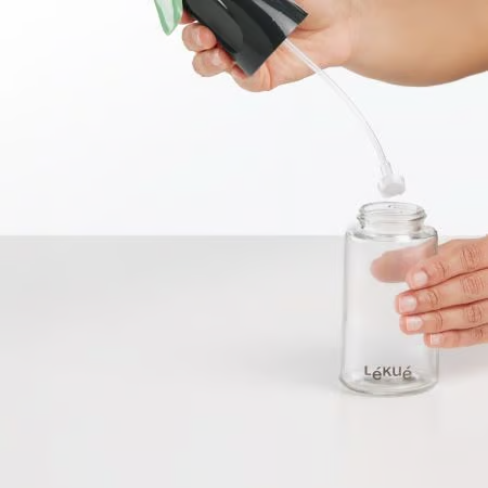 Hand gießt Flüssigkeit aus einer Flasche in eine klare Glasflasche auf weißer Oberfläche.