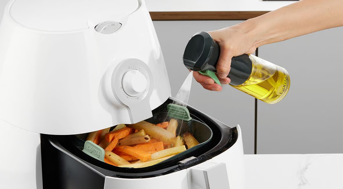 Hand sprüht Öl in eine Heißluftfritteuse mit Essen, auf weißer Arbeitsplatte. Nahaufnahme.