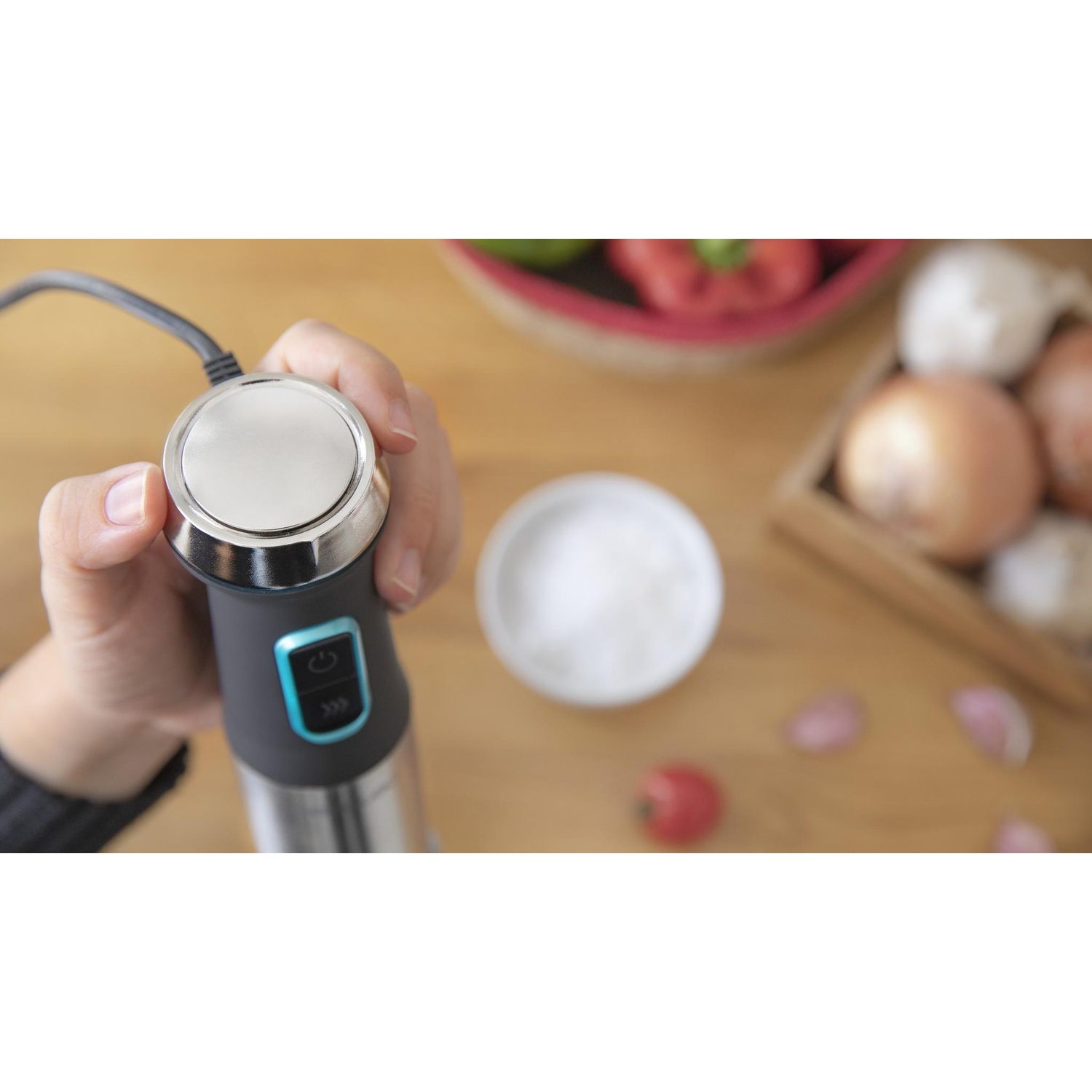 Mano che tiene un frullatore a immersione nero, ingredienti nelle vicinanze.