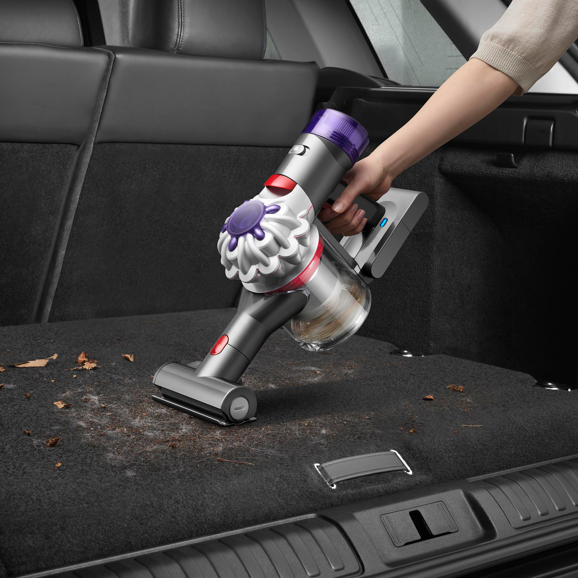 Eine Hand saugt den Kofferraum eines Autos. Der Staubsauger ist grau, lila und rot. Ablagerungen sind sichtbar.
