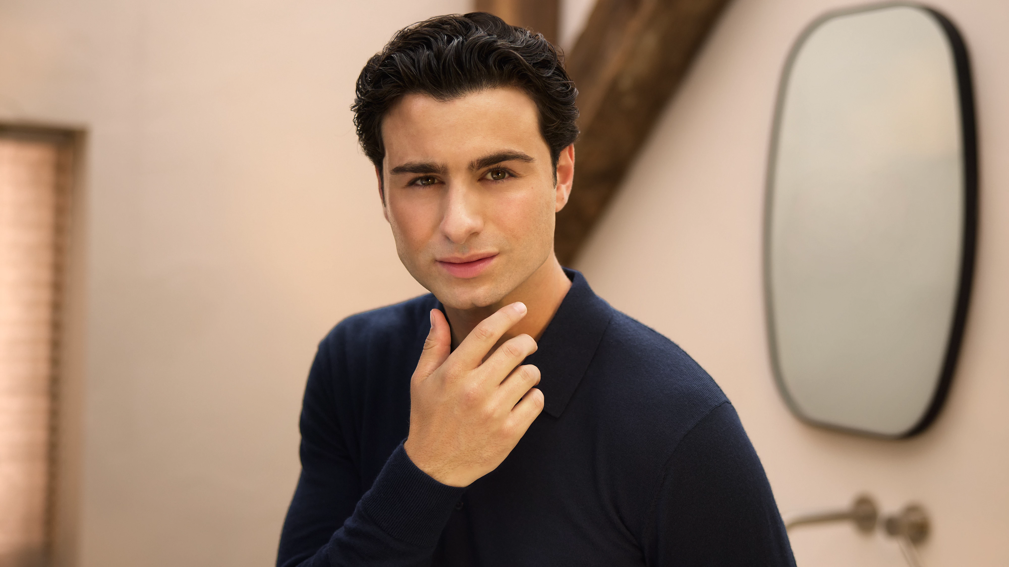 Ein Mann berührt sein Gesicht. Er ist in einem Raum mit einem Spiegel im Hintergrund. Er trägt ein dunkelblaues Hemd.