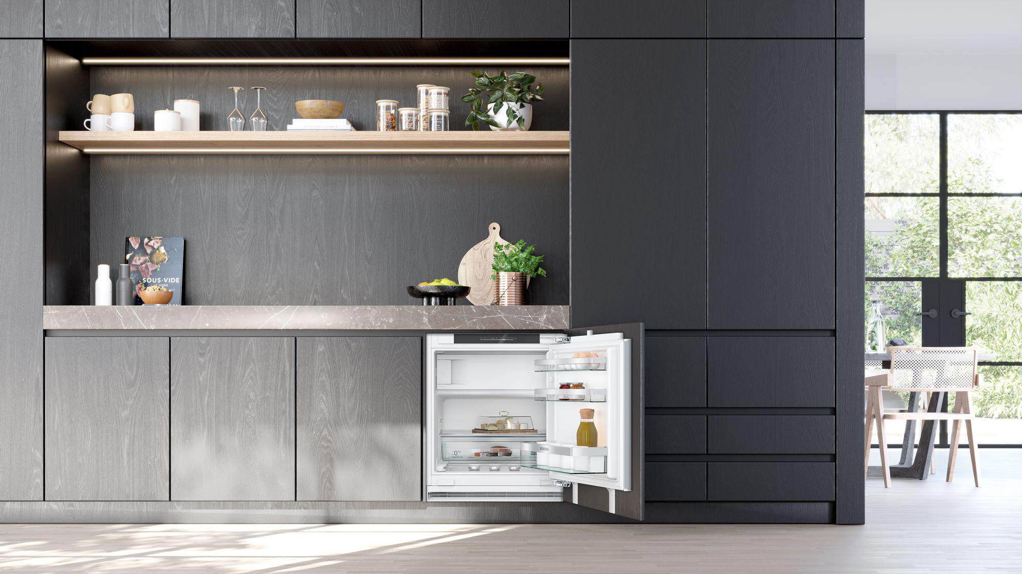 Kücheninterieur mit offenem Kühlschrank. Kühlschrankinhalt: Essen, Getränke. Hölzerne Schränke und Regale im Hintergrund.