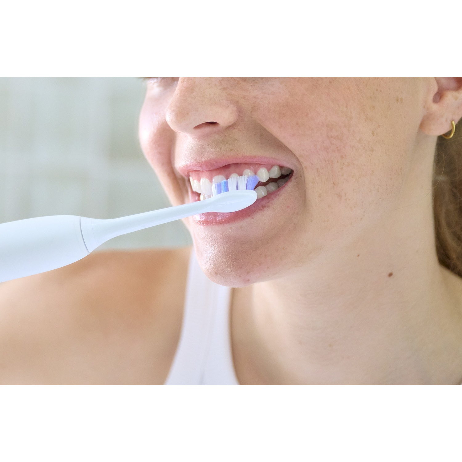 Une femme se brosse les dents avec une brosse à dents électrique blanche. Sa bouche est ouverte.