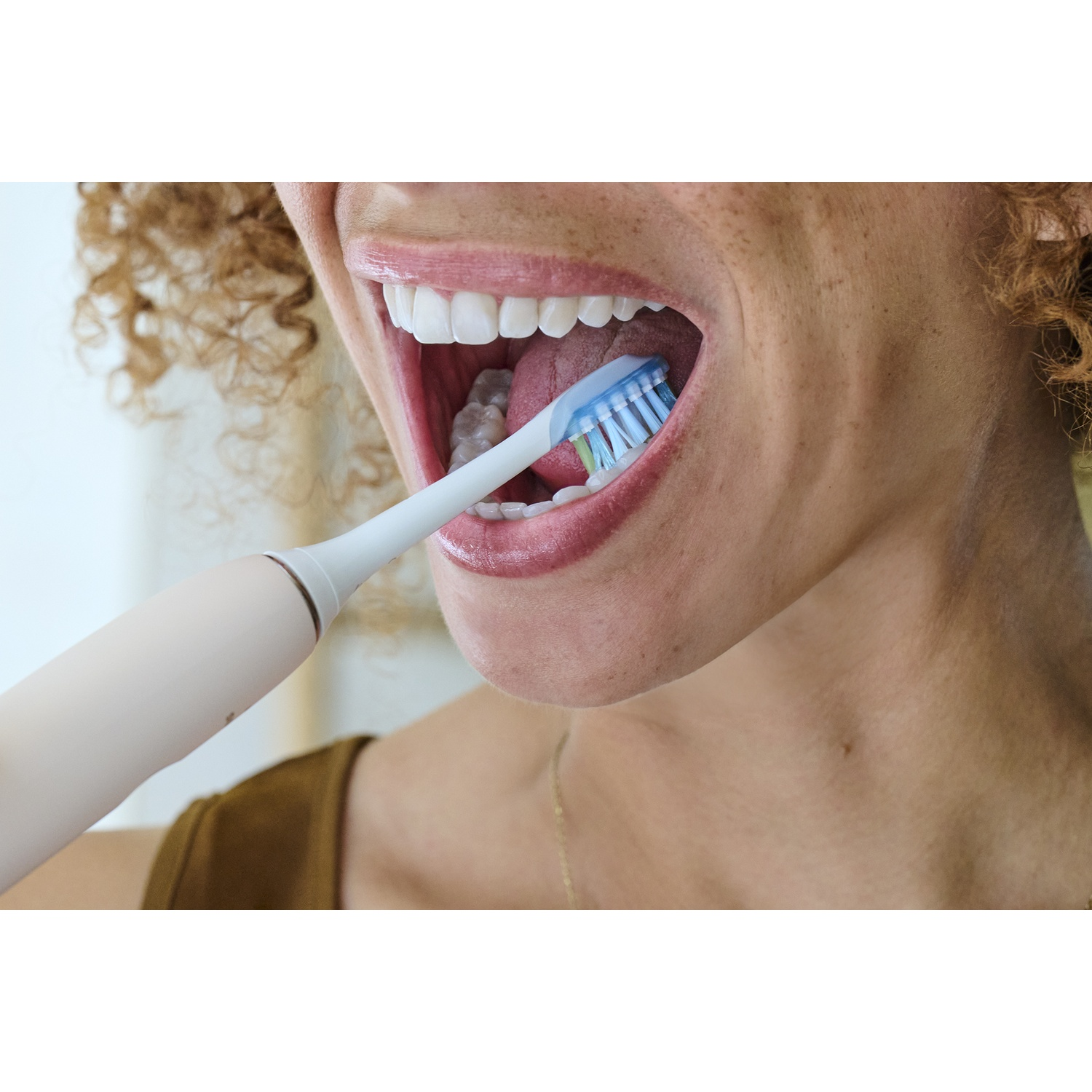 Une femme se brosse les dents avec une brosse à dents électrique. Elle a la bouche ouverte et sourit.