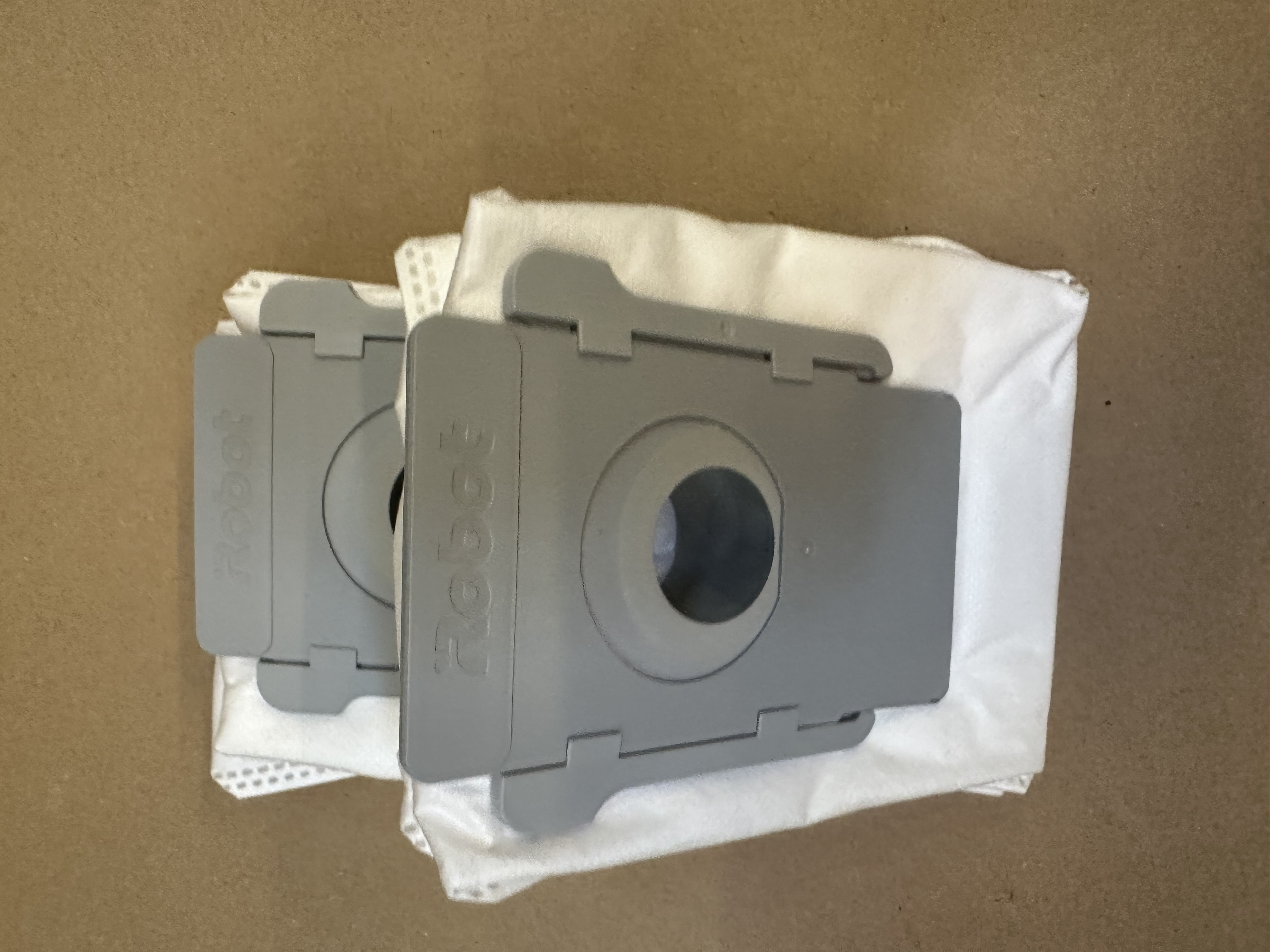 Dwa szare worki do odkurzacza robota, z szarymi plastikowymi pokrywami, leżące na brązowej powierzchni.