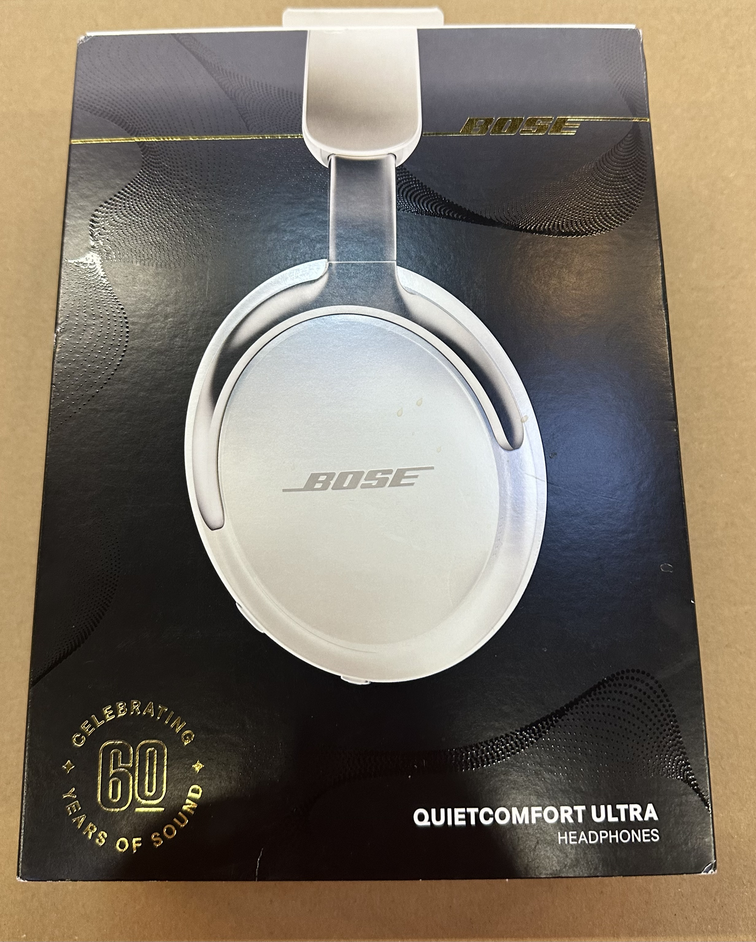 Białe słuchawki Bose w pudełku, czarne i złote akcenty, szczegóły produktu, na stole.