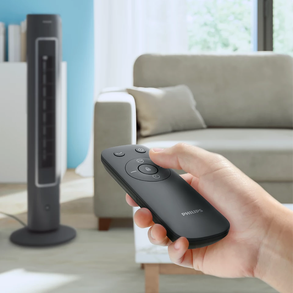 Main tenant une télécommande pour un ventilateur tour gris foncé. Un canapé est en arrière-plan.