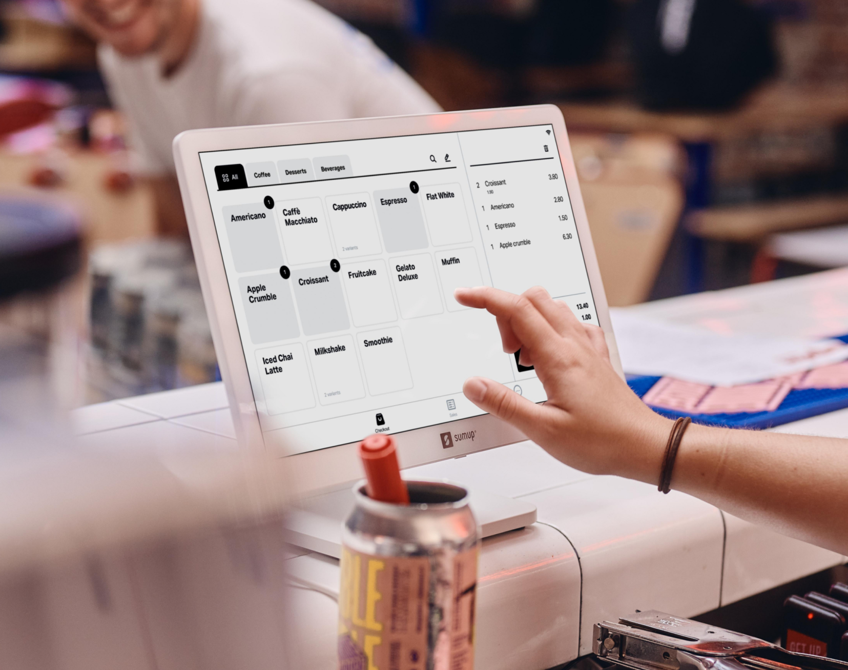 La main d'une personne touche l'écran d'un terminal de point de vente. L'écran affiche un menu.