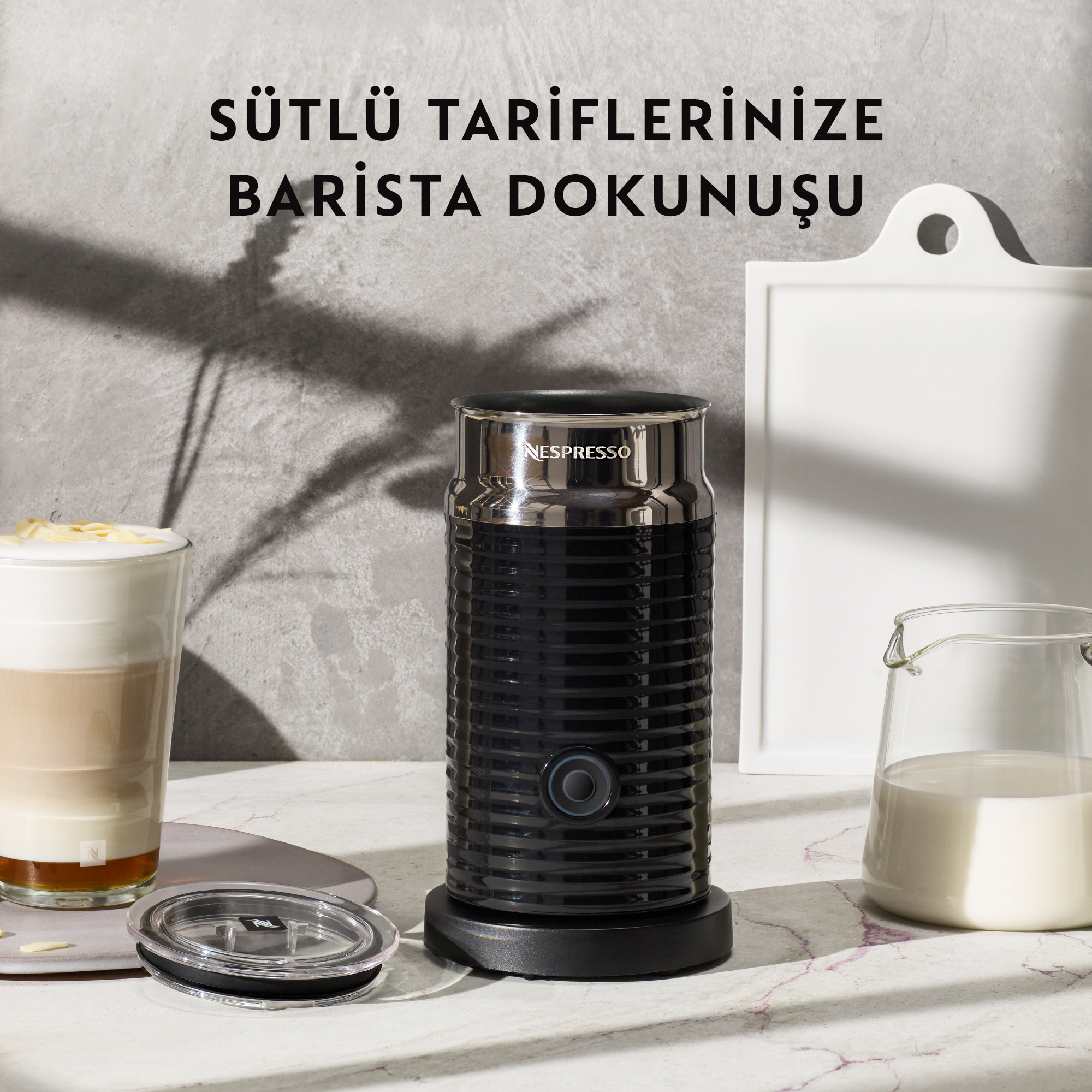 Bir bardak latte, beyaz bir kesme tahtası ve bir süt sürahisi ile siyah bir Nespresso süt köpürtücü.