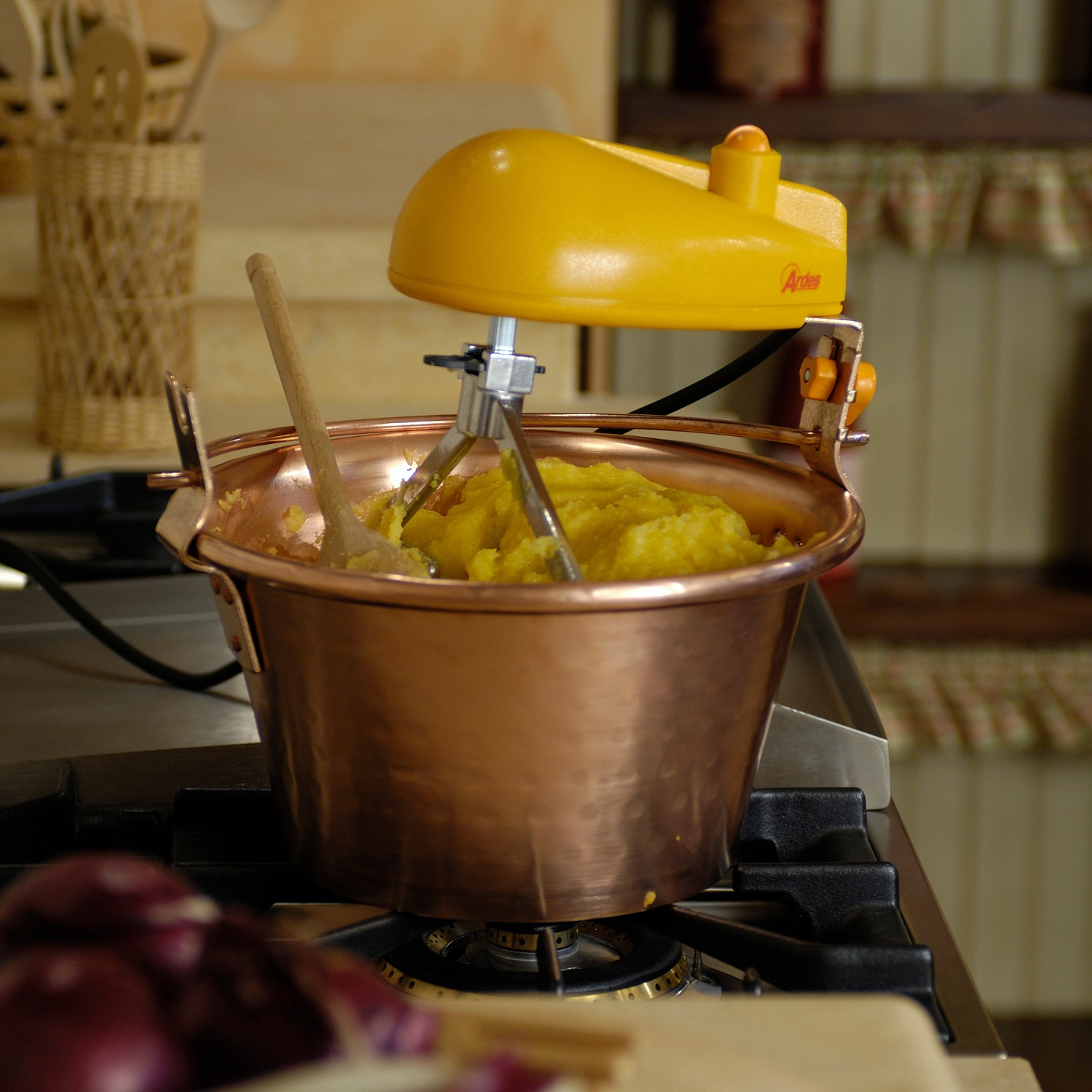 Pentola di rame con composto giallo, cucchiaio di legno, frullatore giallo. Su un fornello. Sfondo scaffali in legno.