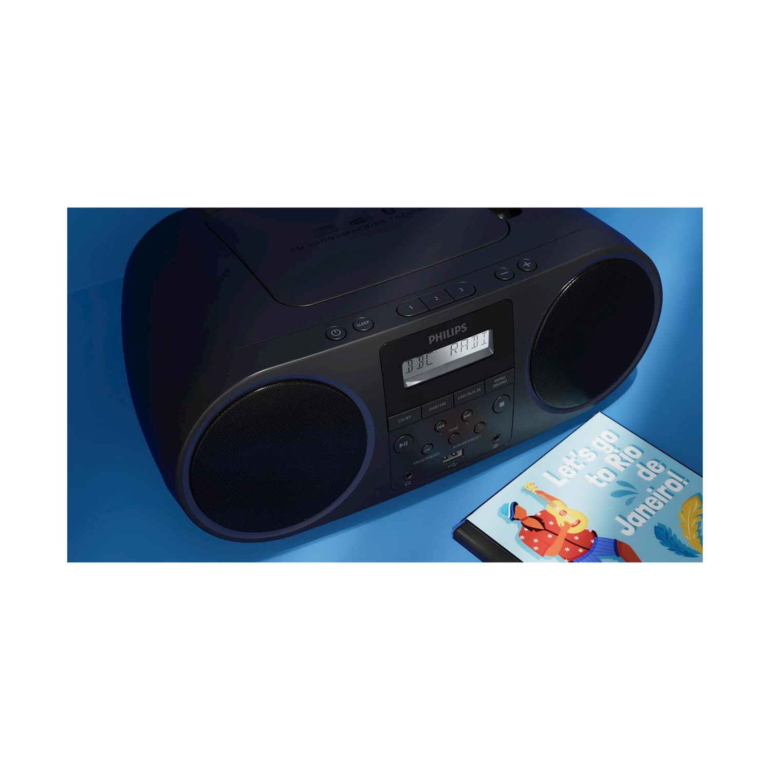 Boombox nera. Marchio Philips, display visibile. Slot CD aperto. Sfondo blu, un libro vicino.