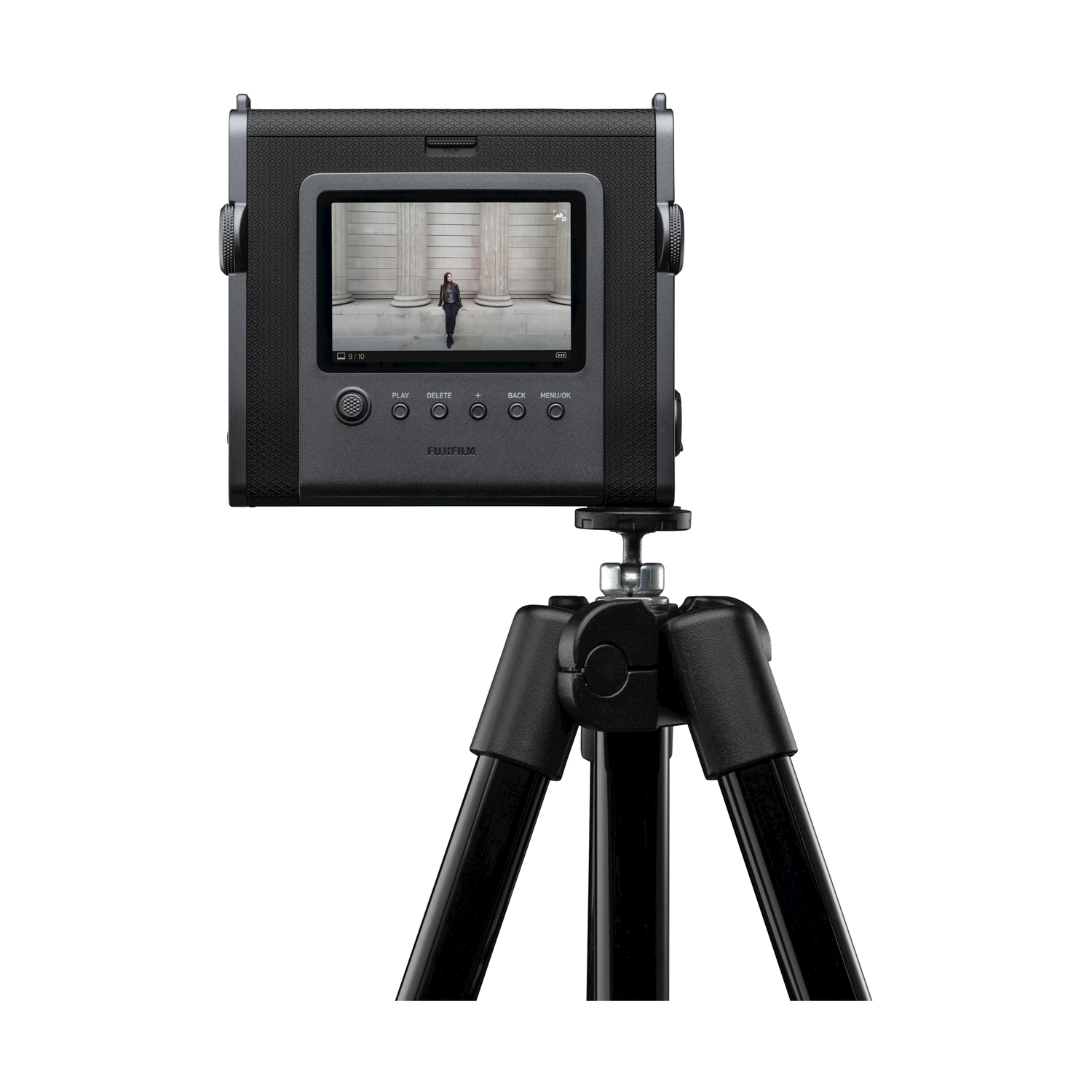 Fotocamera Instax nera su un treppiede, che mostra un'immagine sullo schermo. Sfondo bianco.