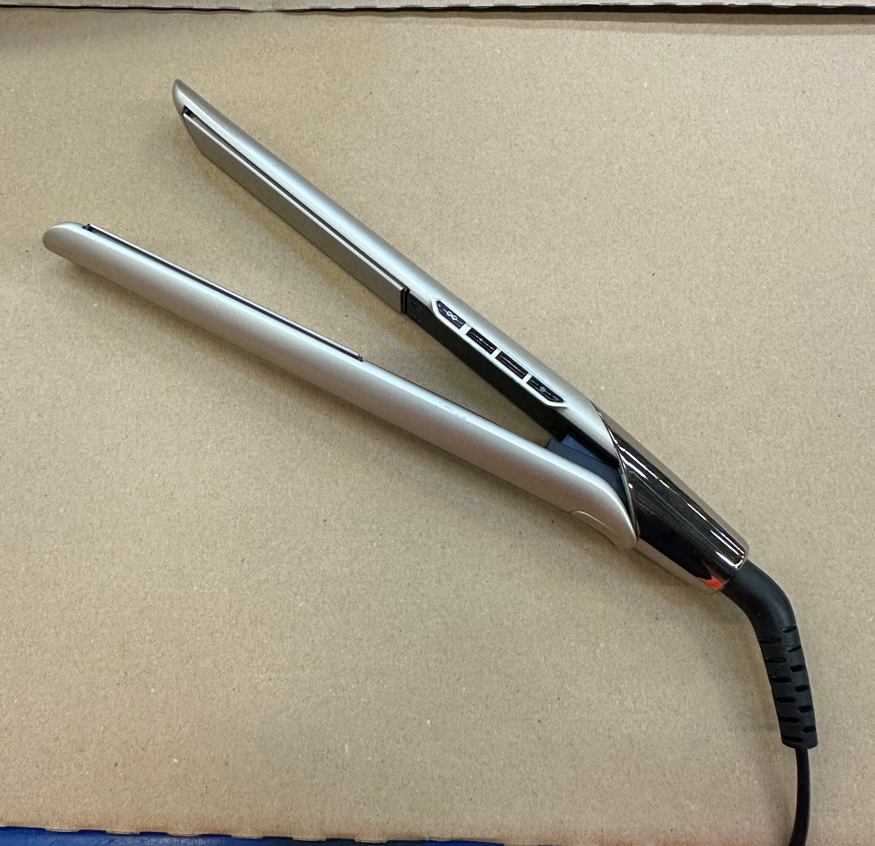 Srebrna prostownica z czarnym kablem leży na brązowej powierzchni.