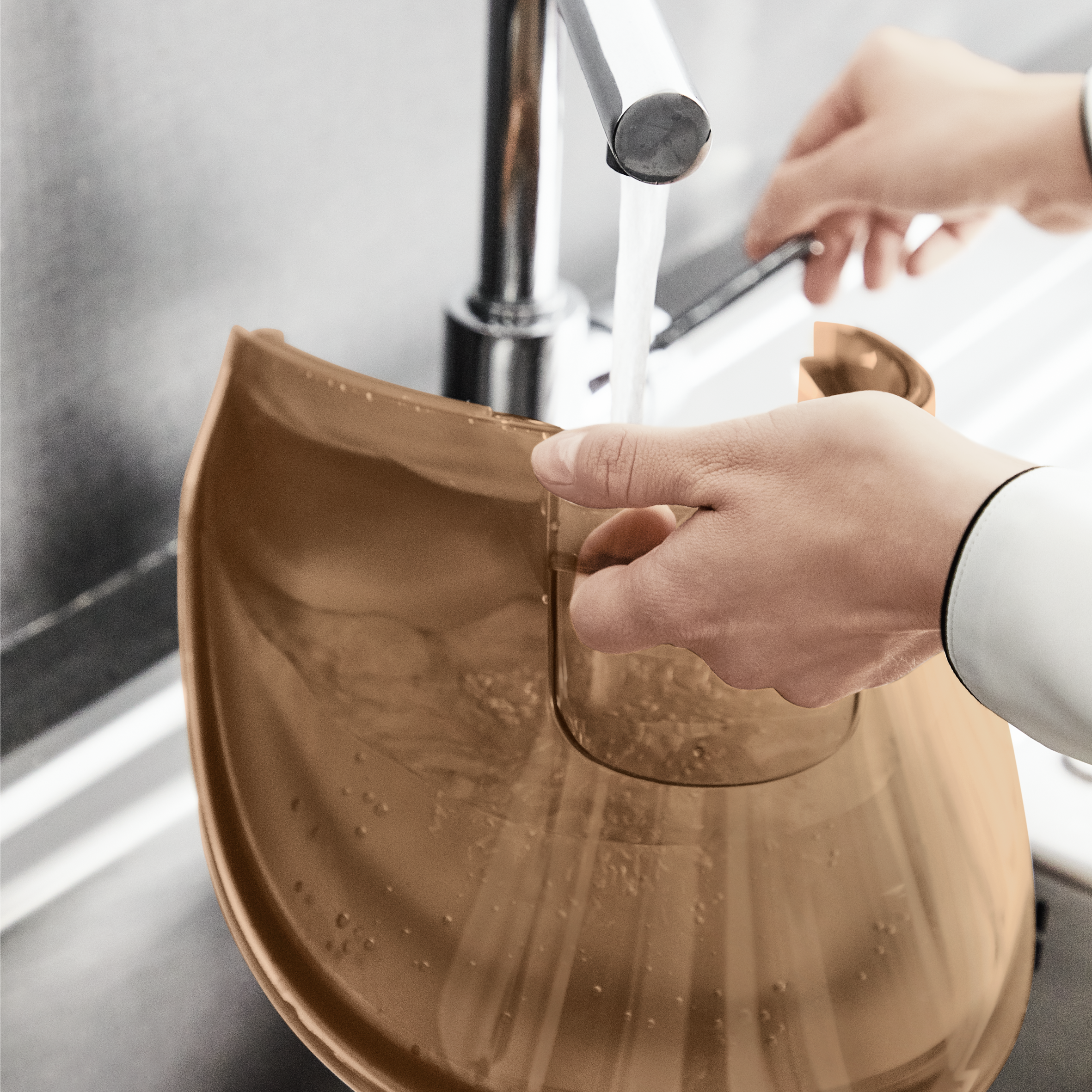 Woda wlewana do brązowego pojemnika w zlewie, widoczne dłonie osoby.