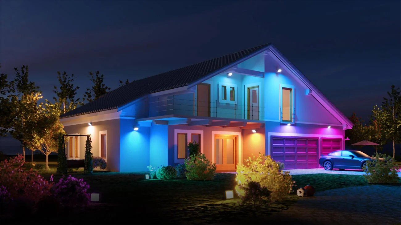 Dom w nocy, oświetlony kolorowymi światłami. Samochód na podjeździe. Ogród z krzewami.