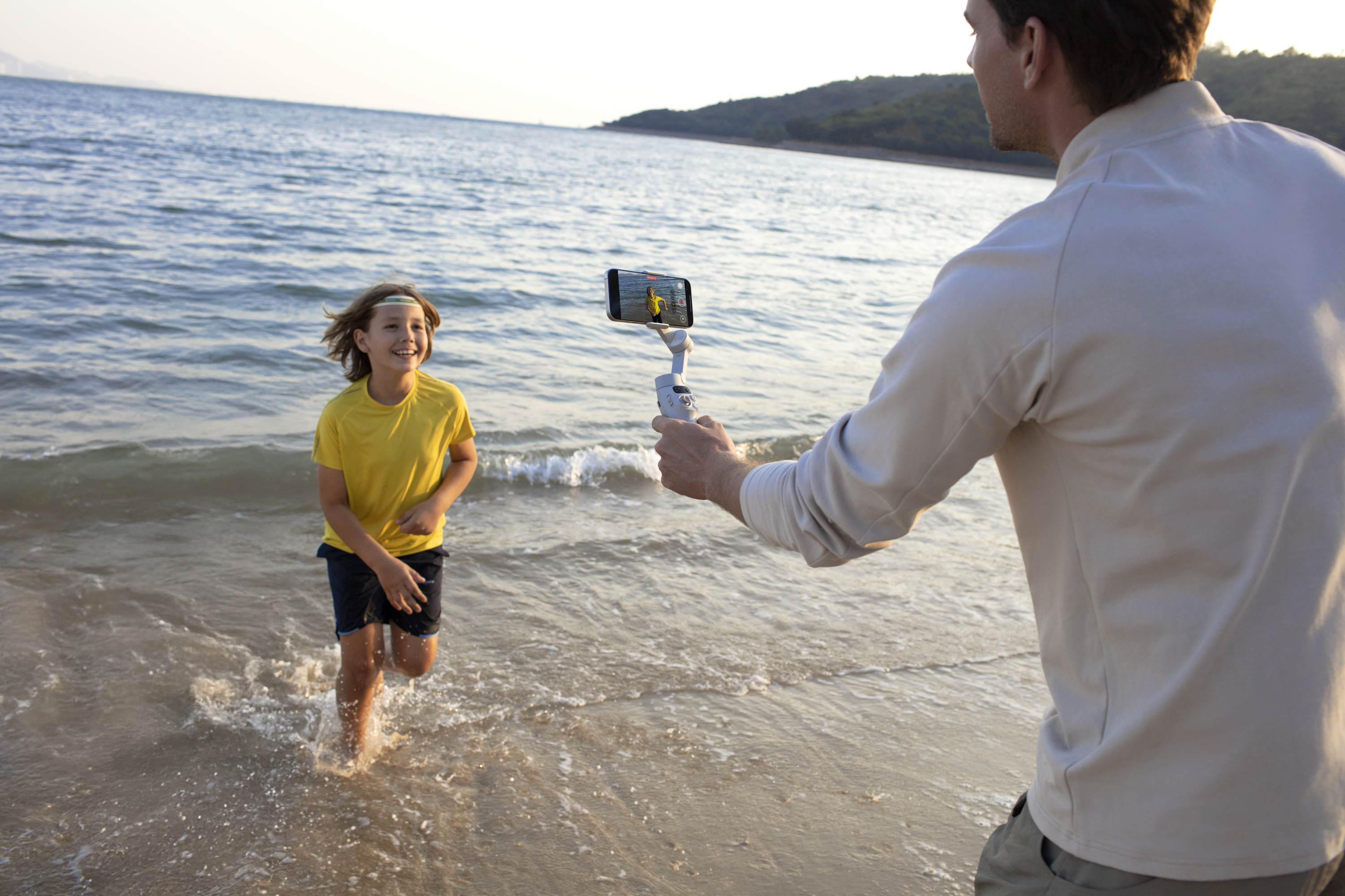 Un homme filme un enfant courant sur une plage. L'enfant sourit.