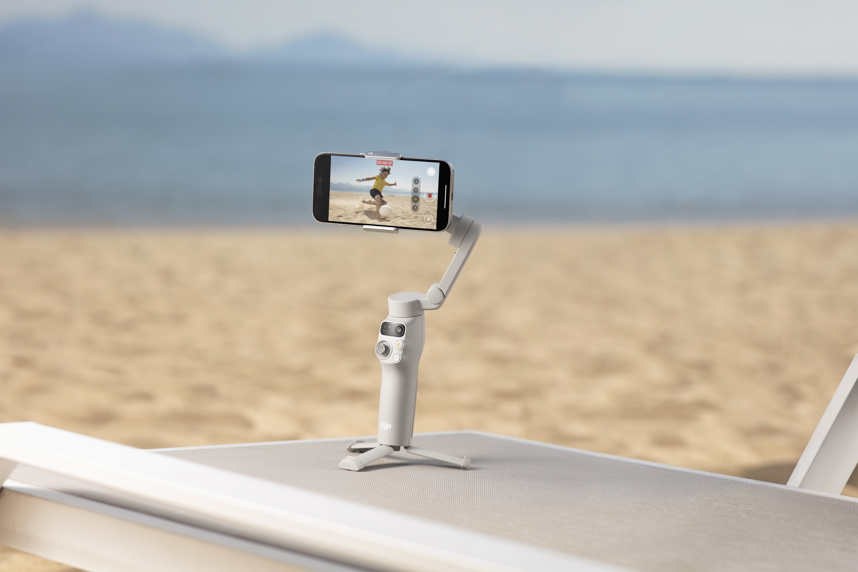 Un cardan blanc tient un téléphone enregistrant une personne sur une plage. L'arrière-plan est une plage et un océan flous.