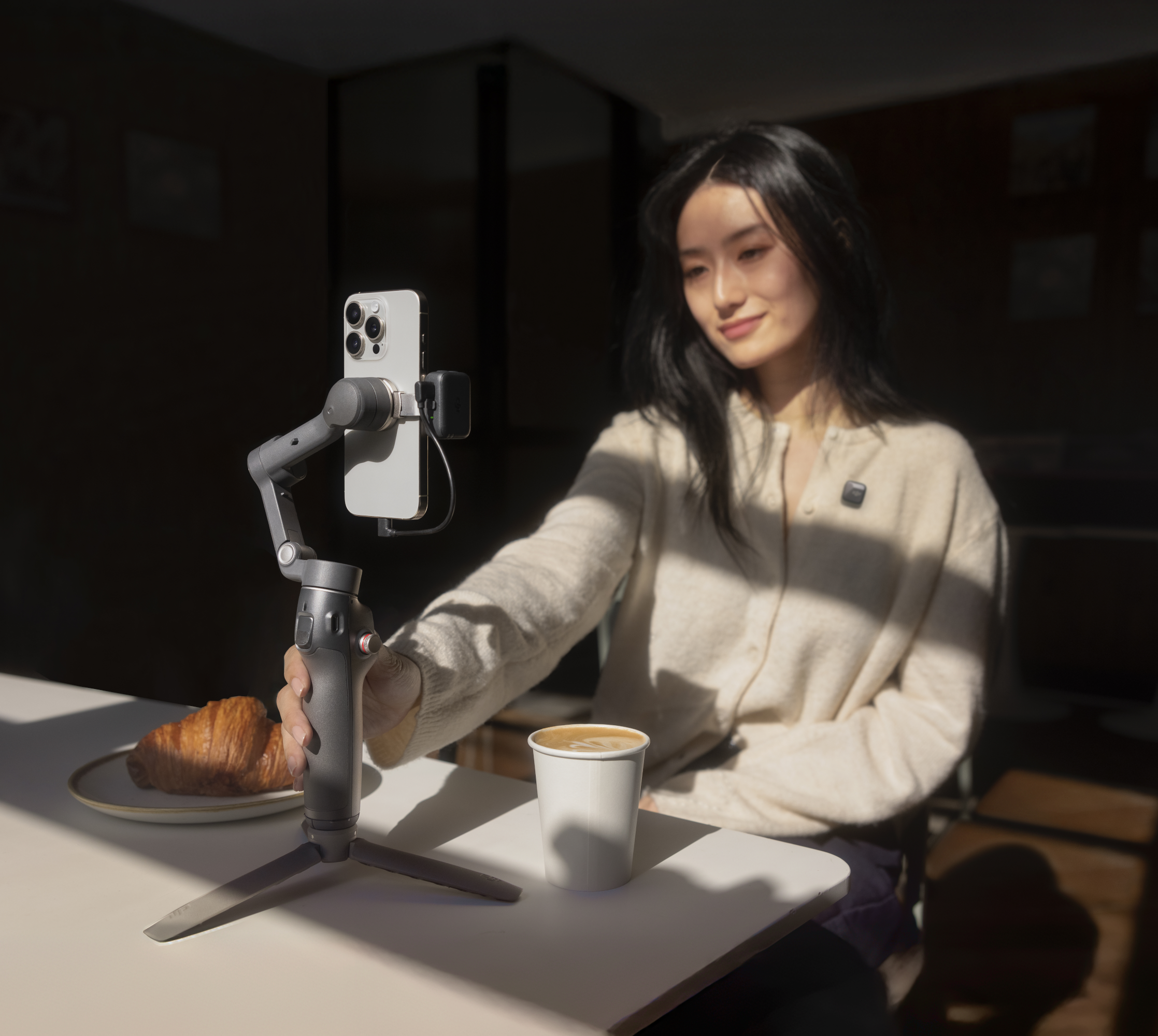 Femme tenant un téléphone sur un support, croissant et tasse à café sur la table.