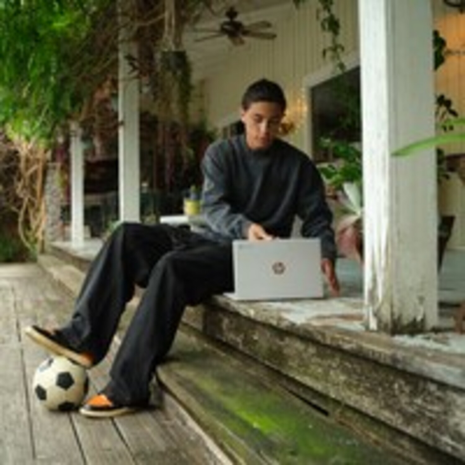 A young man is sitting on steps outside using a laptop with a soccer ball nearby.