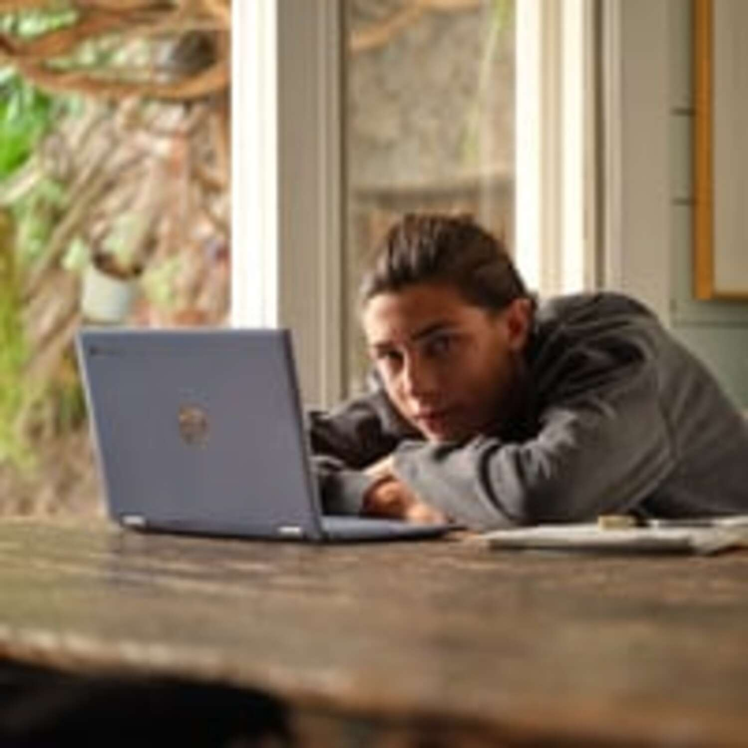 A person is using a blue laptop. They are leaning on a wooden table. The window is on the background.