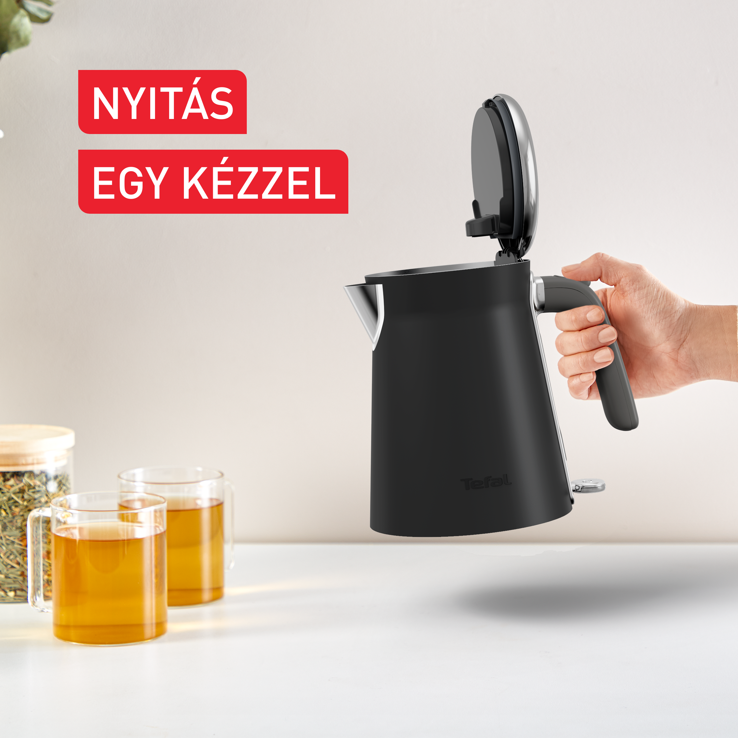 Egy fekete vízforraló nyitott fedéllel, egy kéz tartja. A szöveg: 'Nyitás egy kézzel'.