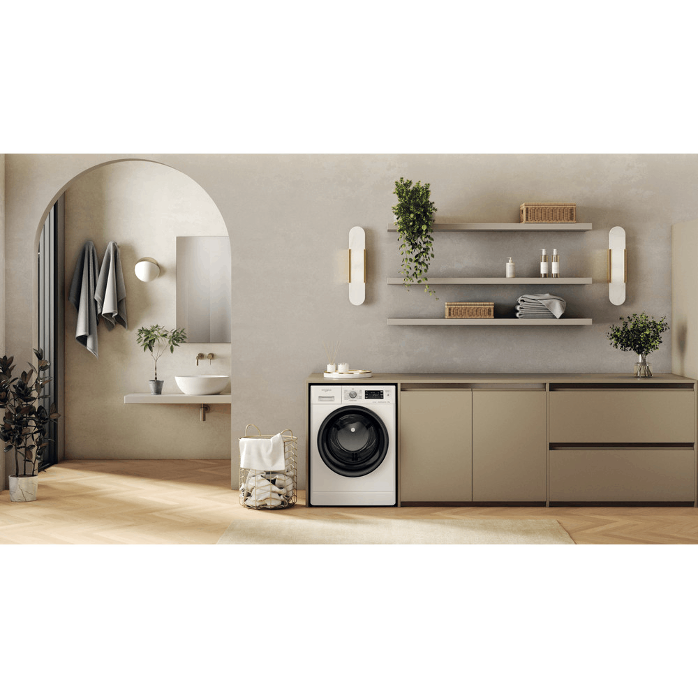 Lavadora blanca en un baño beige con pisos de madera, estantes y un espejo. Hay plantas y decoración.
