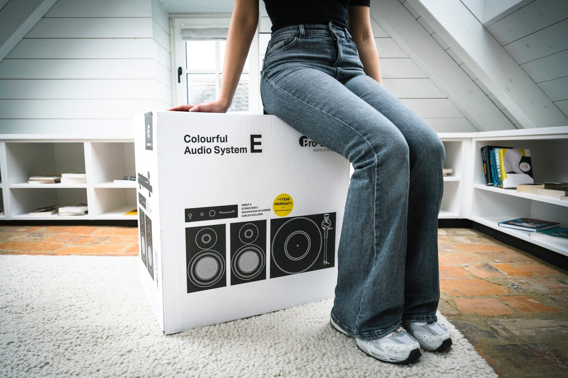 Eine Frau sitzt auf einer bunten Audio-System-Box auf einem weißen Teppich, mit weißem Hintergrund.