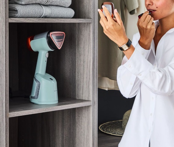 Kobieta w białej koszuli nakłada szminkę w szafie, z parownicą na półce. Widoczne ubrania.