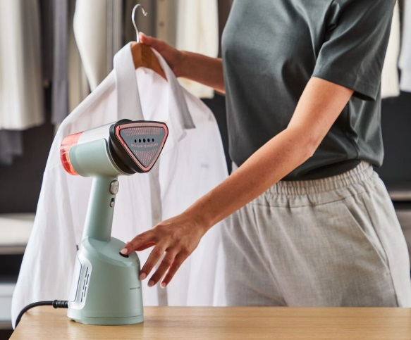 Kobieta trzyma koszulę na wieszaku, używając parownicy ręcznej. Drewniany stół na pierwszym planie.