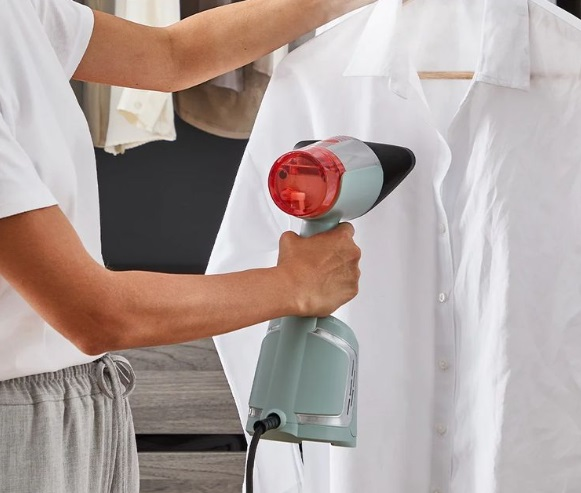 Kobieta prasuje białą koszulę parownicą ręczną. Ubrania wiszące w tle są lekko rozmyte.