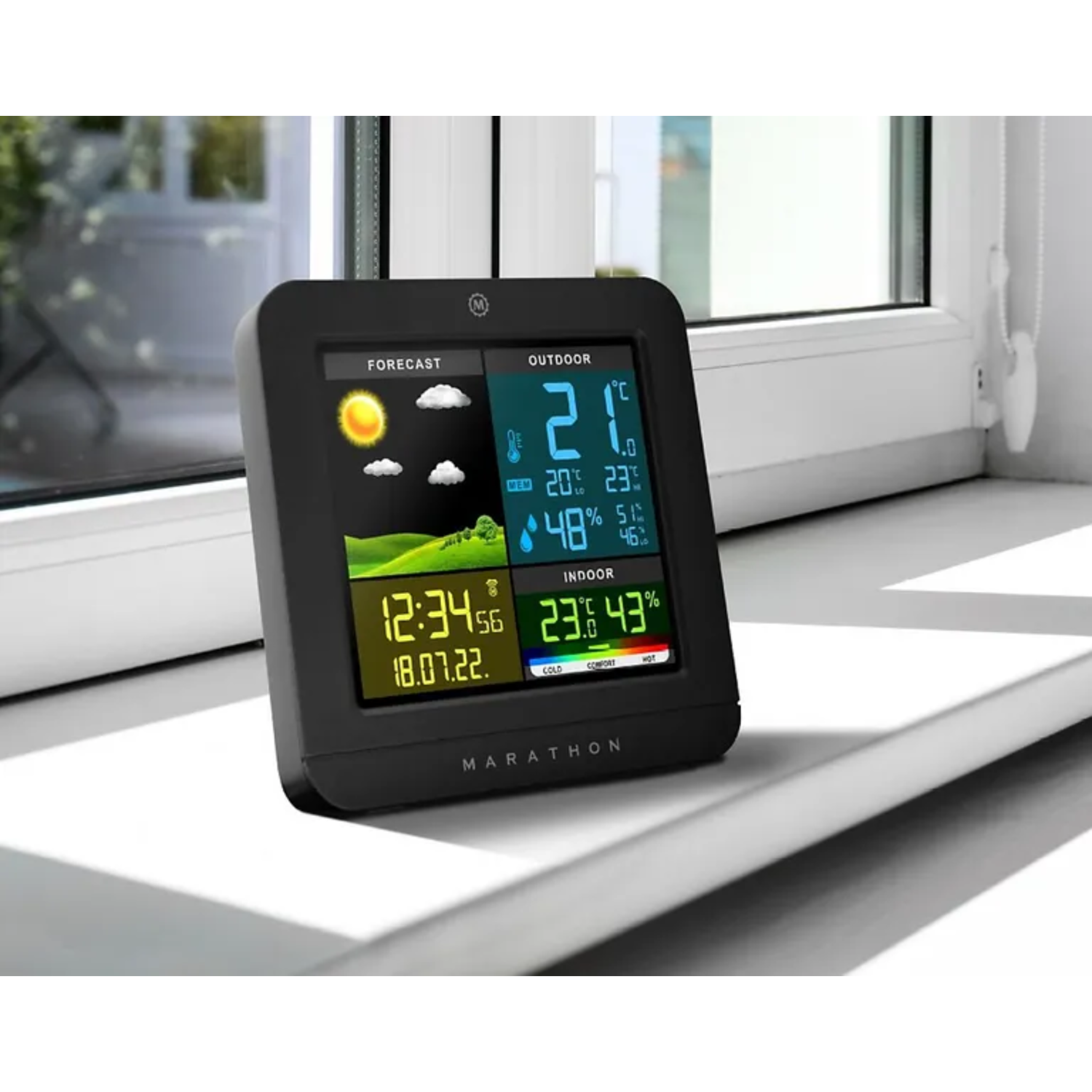A black weather station on a window sill. It displays temperature, forecast, time, and other weather data.