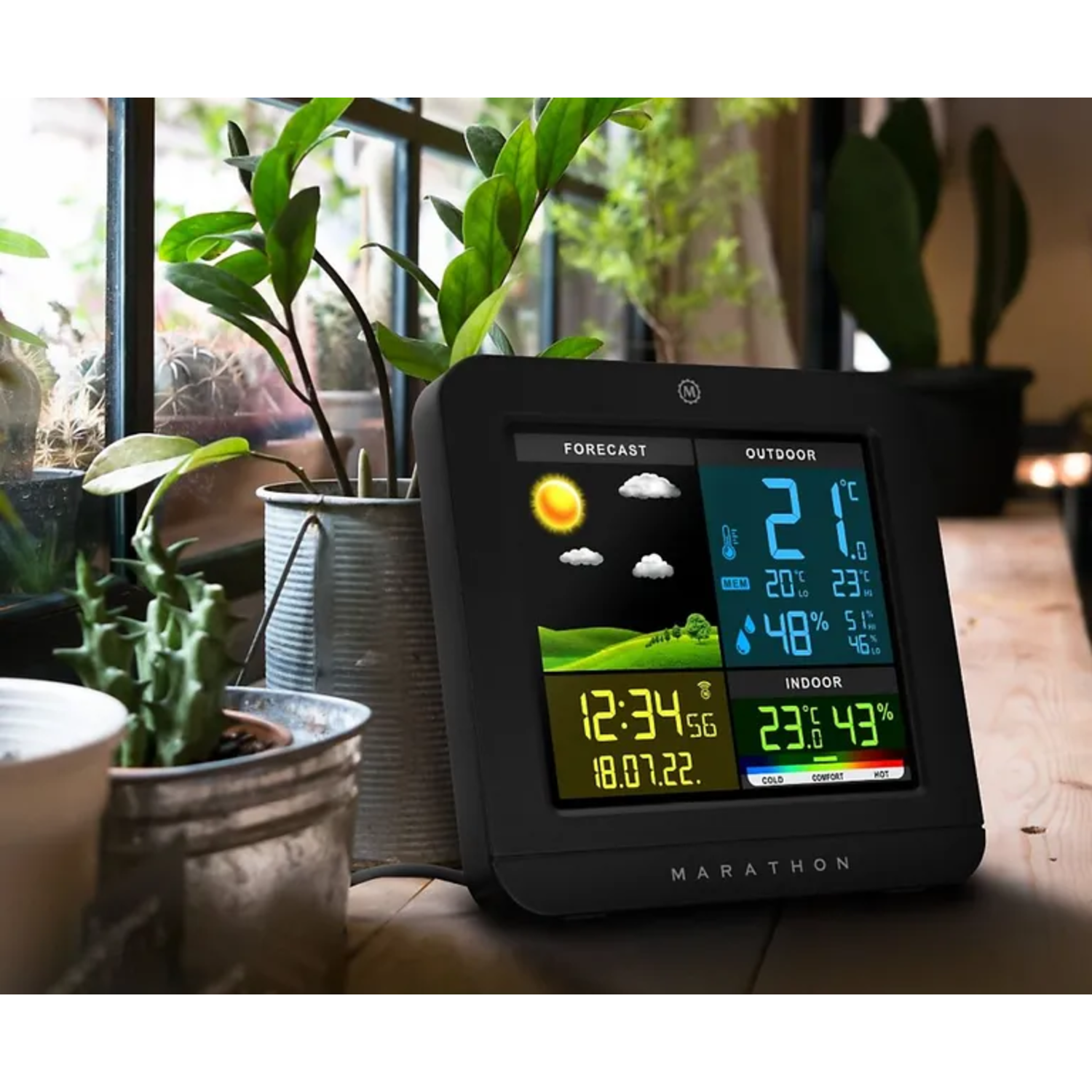 Black weather station displaying forecast, outdoor, and indoor conditions, with plants in the background.