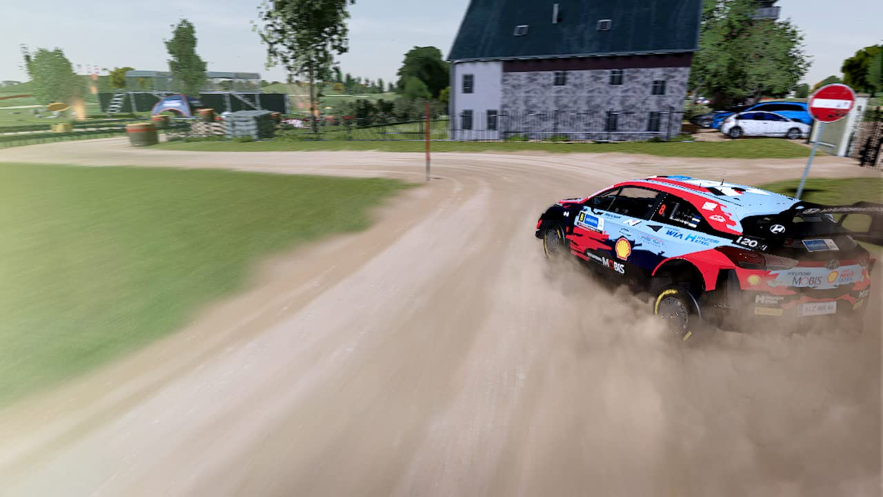 Ein Rallyeauto driftet auf einer unbefestigten Straße und wirbelt Staub auf. Grüne Felder und Gebäude im Hintergrund.