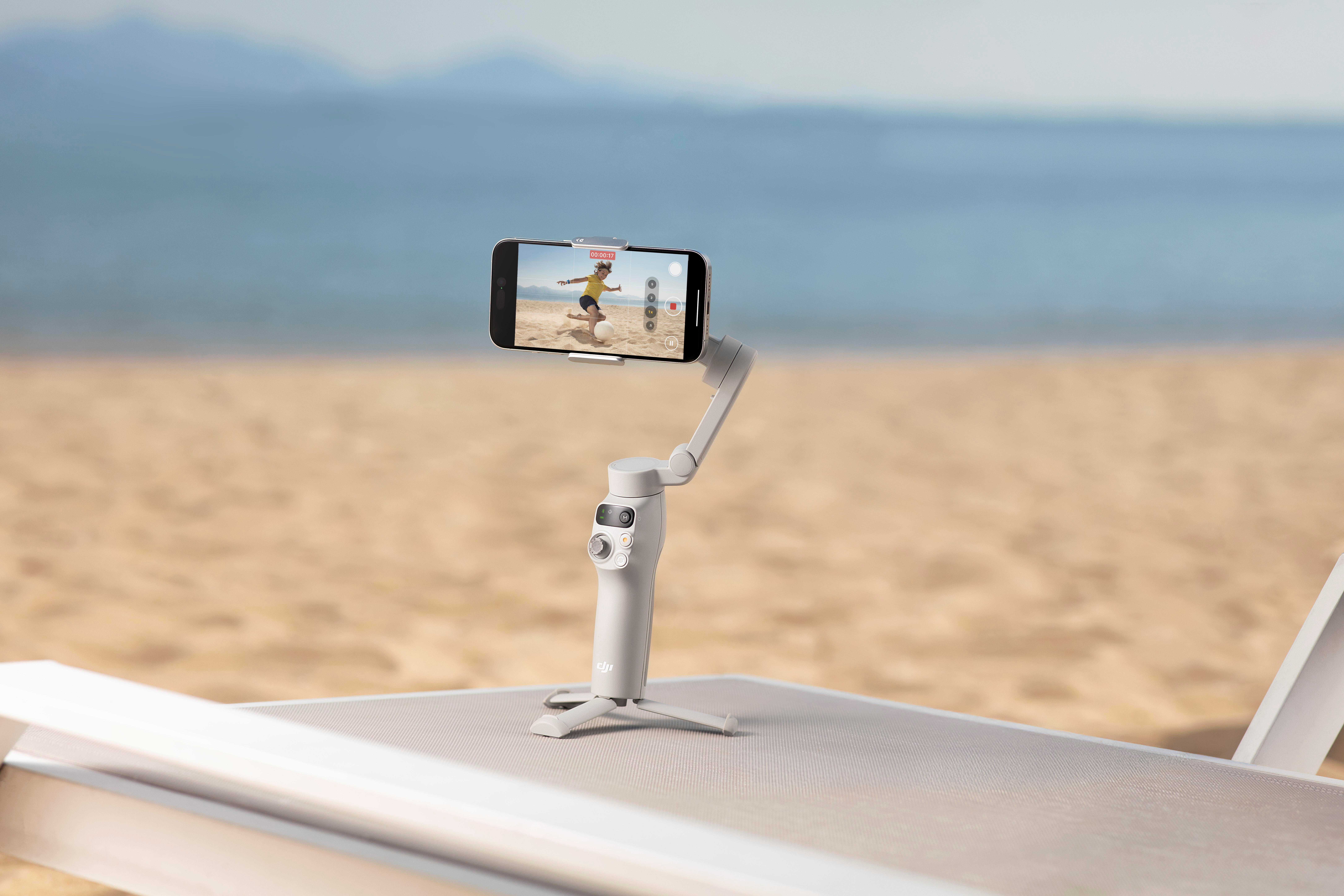 Smartphone auf einem weißen Gimbal, der eine Person am Strand aufnimmt. Sandiger Hintergrund.