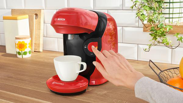 Eine rote Kaffeemaschine mit einer weißen Tasse, eine Hand drückt den Knopf.