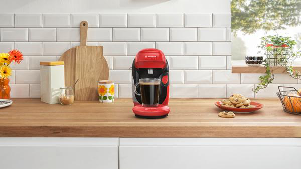 Rote Kaffeemaschine mit einer Tasse, Keksen und Blumen auf einer Holz-Arbeitsplatte in einer Küche.