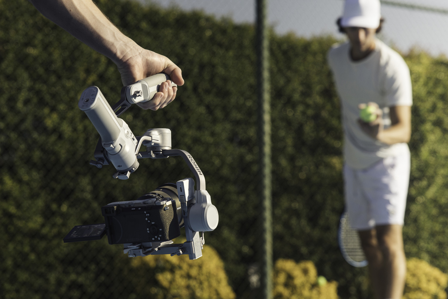 Ręka trzymająca stabilizator kamery, tenisista w tle.