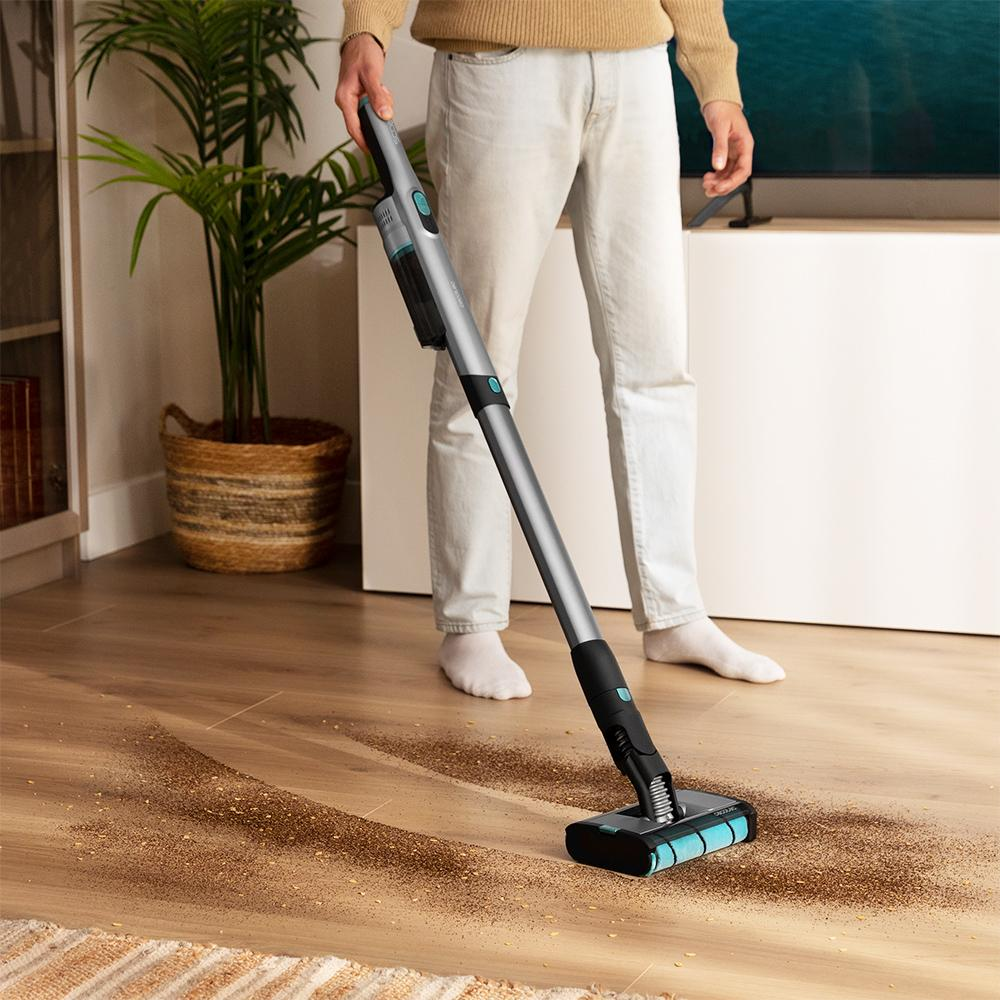 Hombre limpiando el suelo con una aspiradora. Suelo de madera, planta y alfombra visibles.