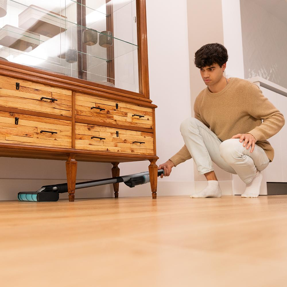 Un hombre se agacha, aspirando debajo de un mueble de madera en un suelo de madera.