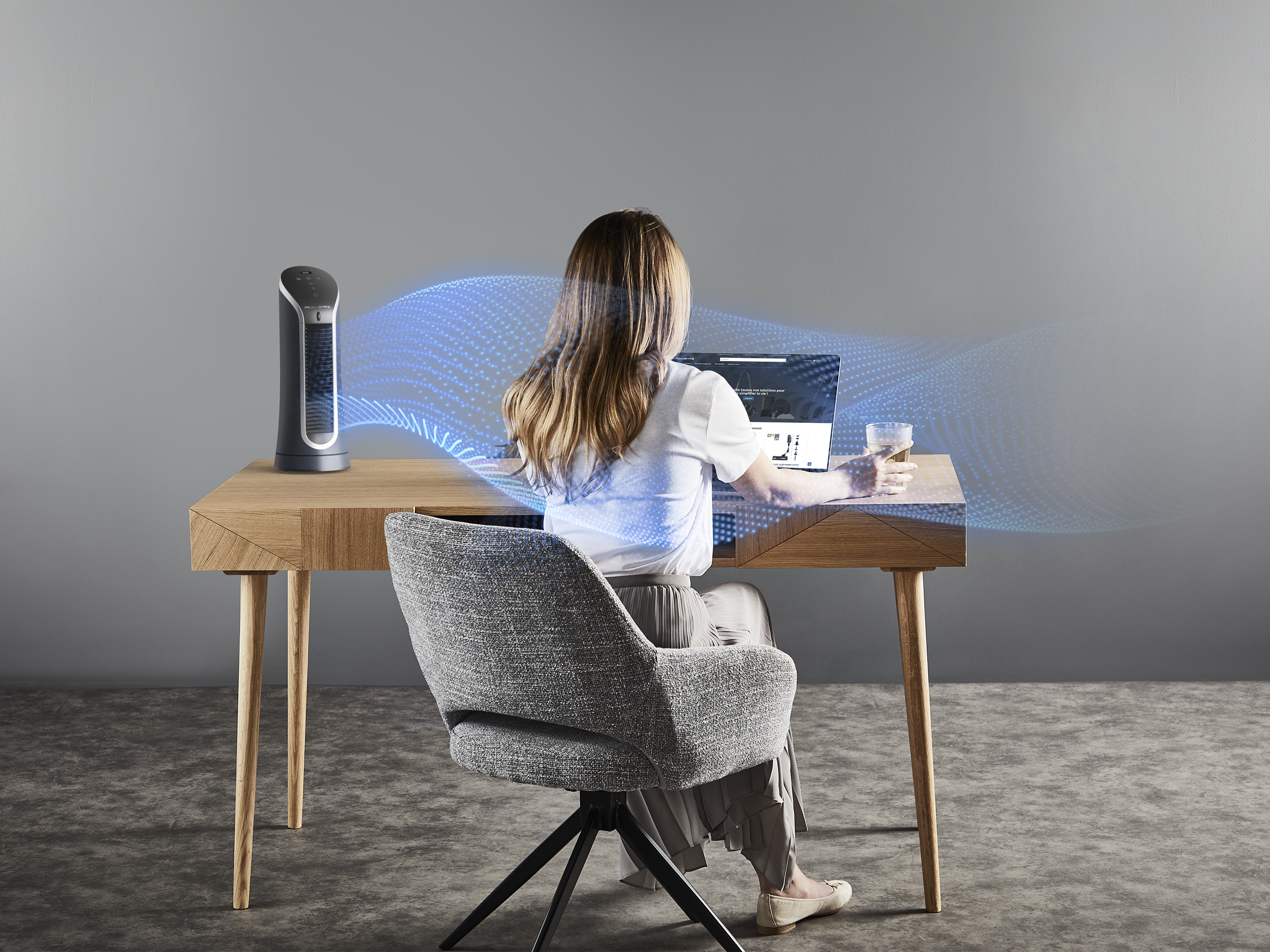 Nő asztalnál laptoppal, ventilátor fújja a levegőt felé.