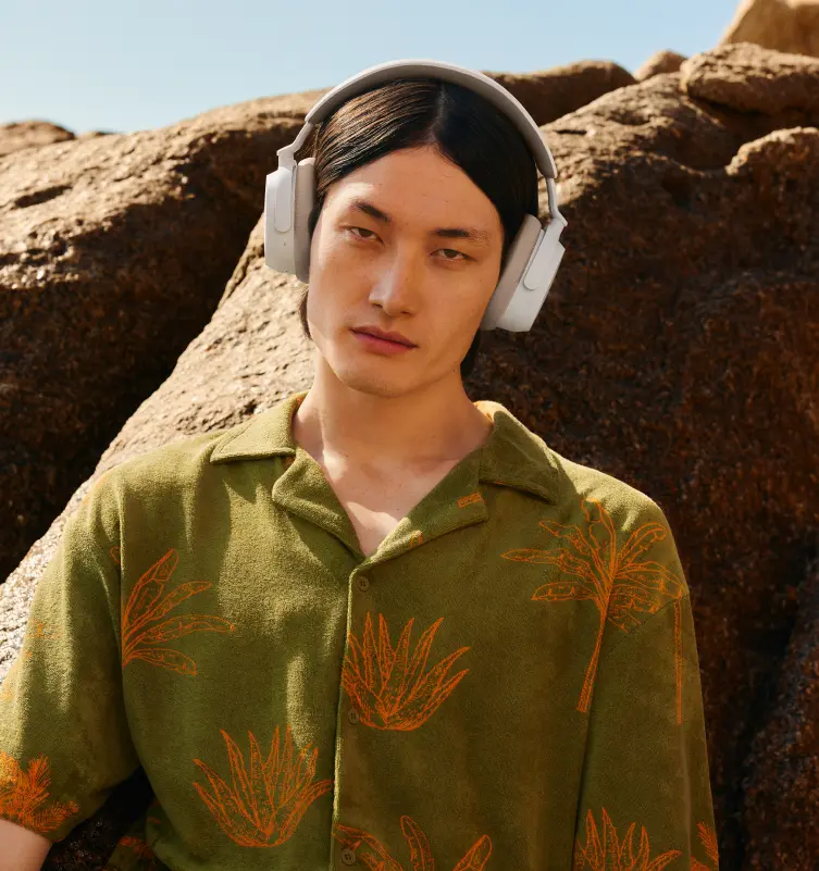 Uomo con cuffie bianche, camicia verde con palme arancioni, in piedi vicino a rocce.