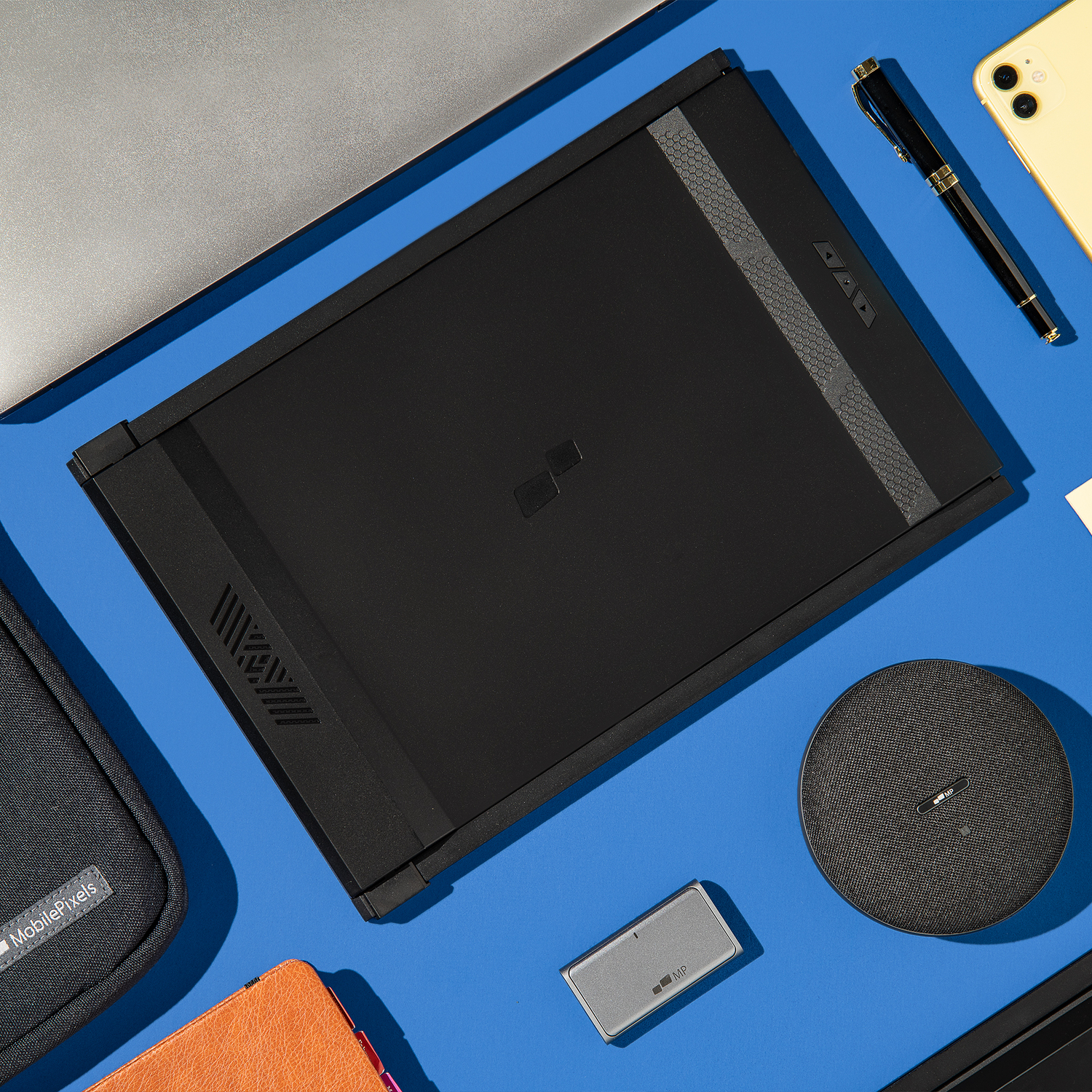 Un ordinateur portable noir est ouvert sur une surface bleue. Un téléphone jaune, un stylo et d'autres objets sont à proximité.