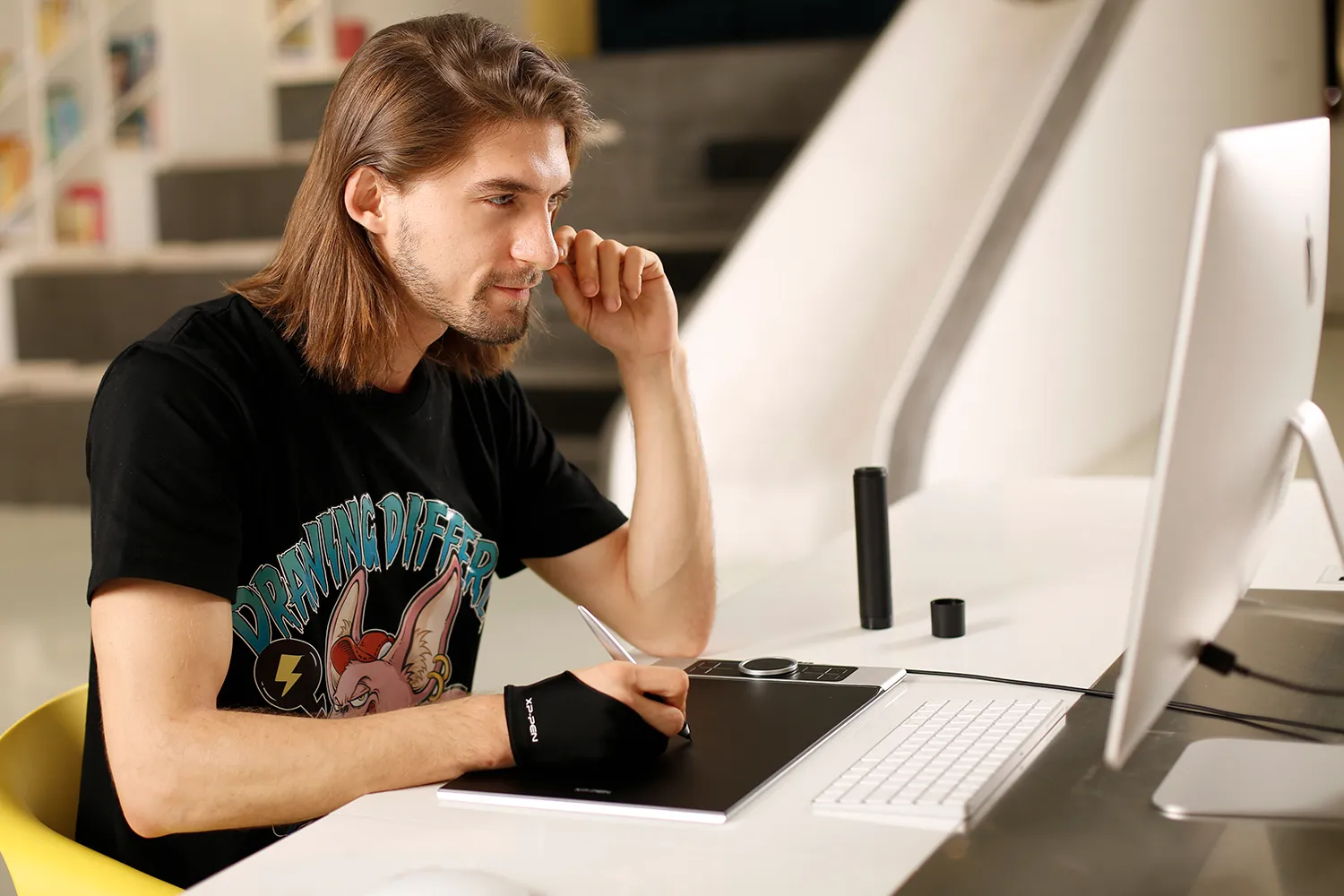 Hosszú hajú férfi XP-Pen táblagépet használ, ül. A számítógép monitorok jobbra. Fekete póló.