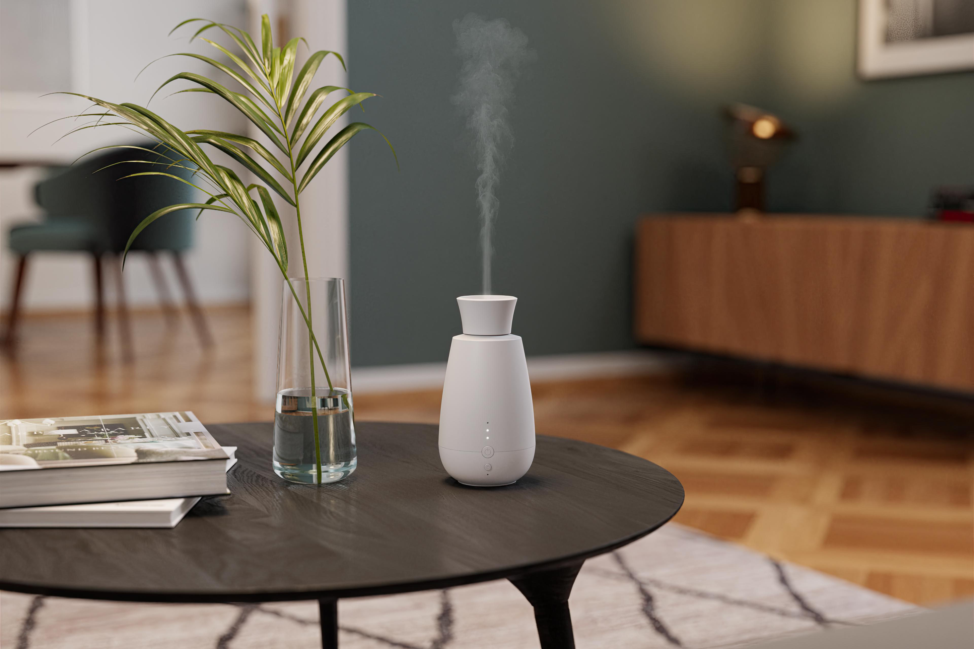 Weißer Diffusor für ätherische Öle, der Dampf abgibt, mit Pflanze und Büchern auf Tisch.