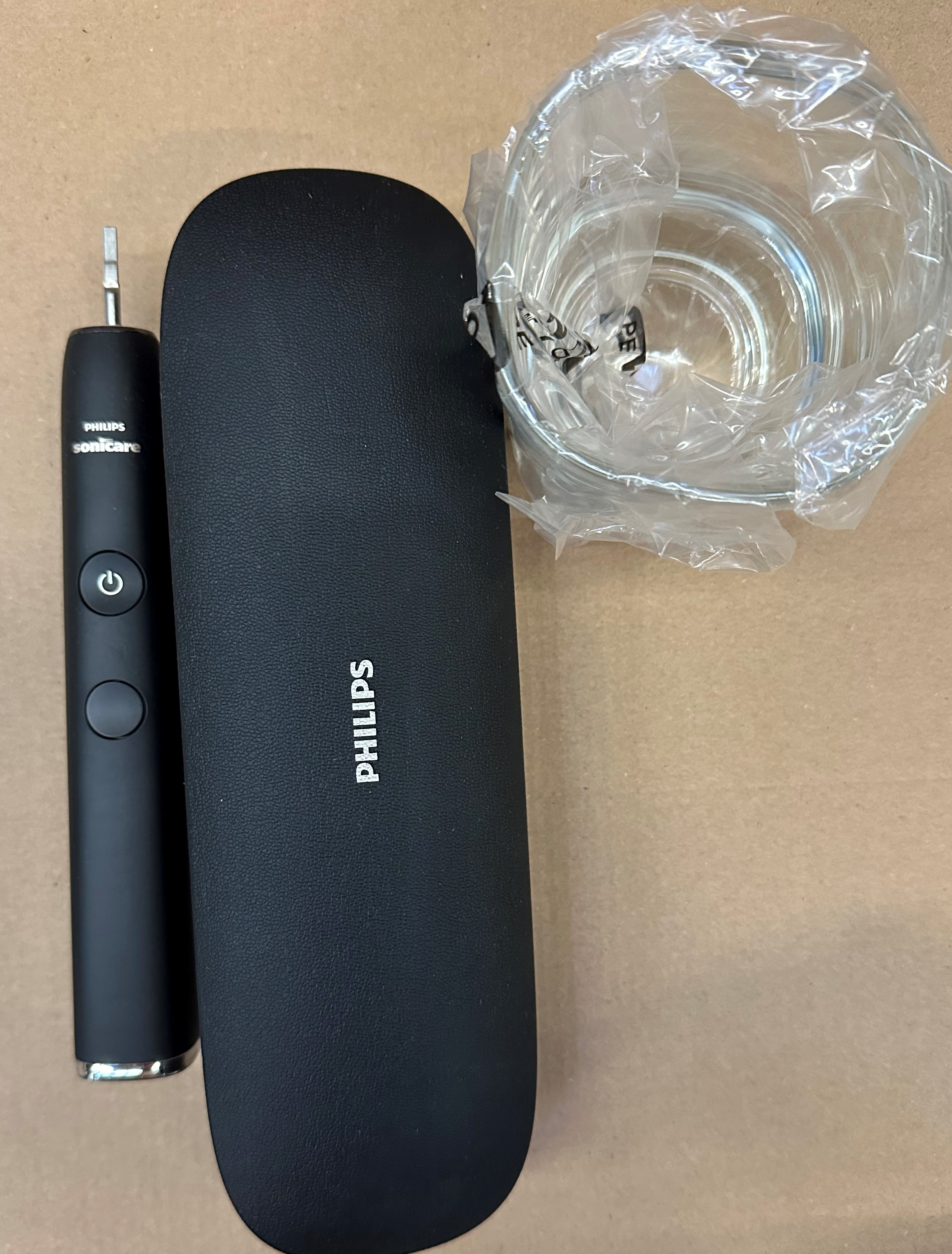 Czarna szczoteczka elektryczna, etui i szklanka w folii na brązowym tle.