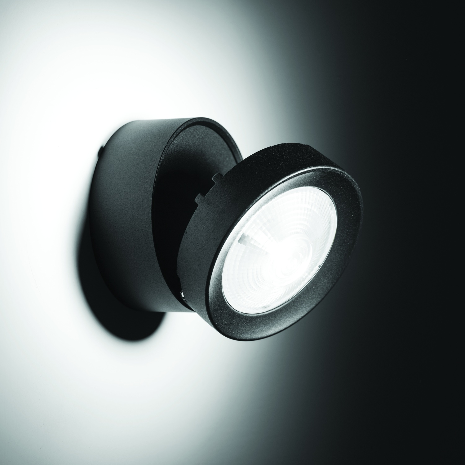 Faretto nero montato a parete, angolato verso lo spettatore, illuminato e che proietta un'ombra.