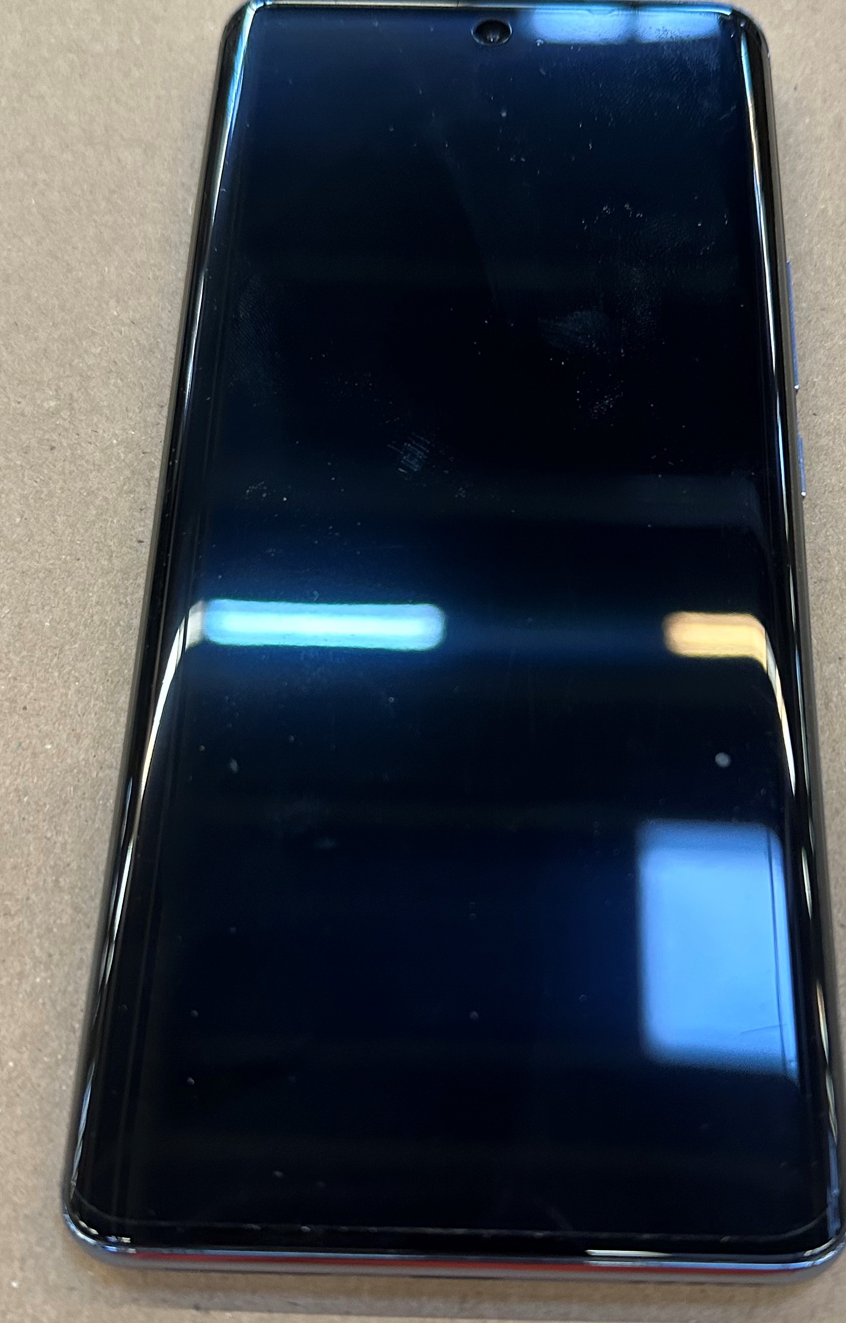 Czarny smartfon pod kątem na brązowej powierzchni. Ekran jest czarny i błyszczący. Telefon ma aparat.