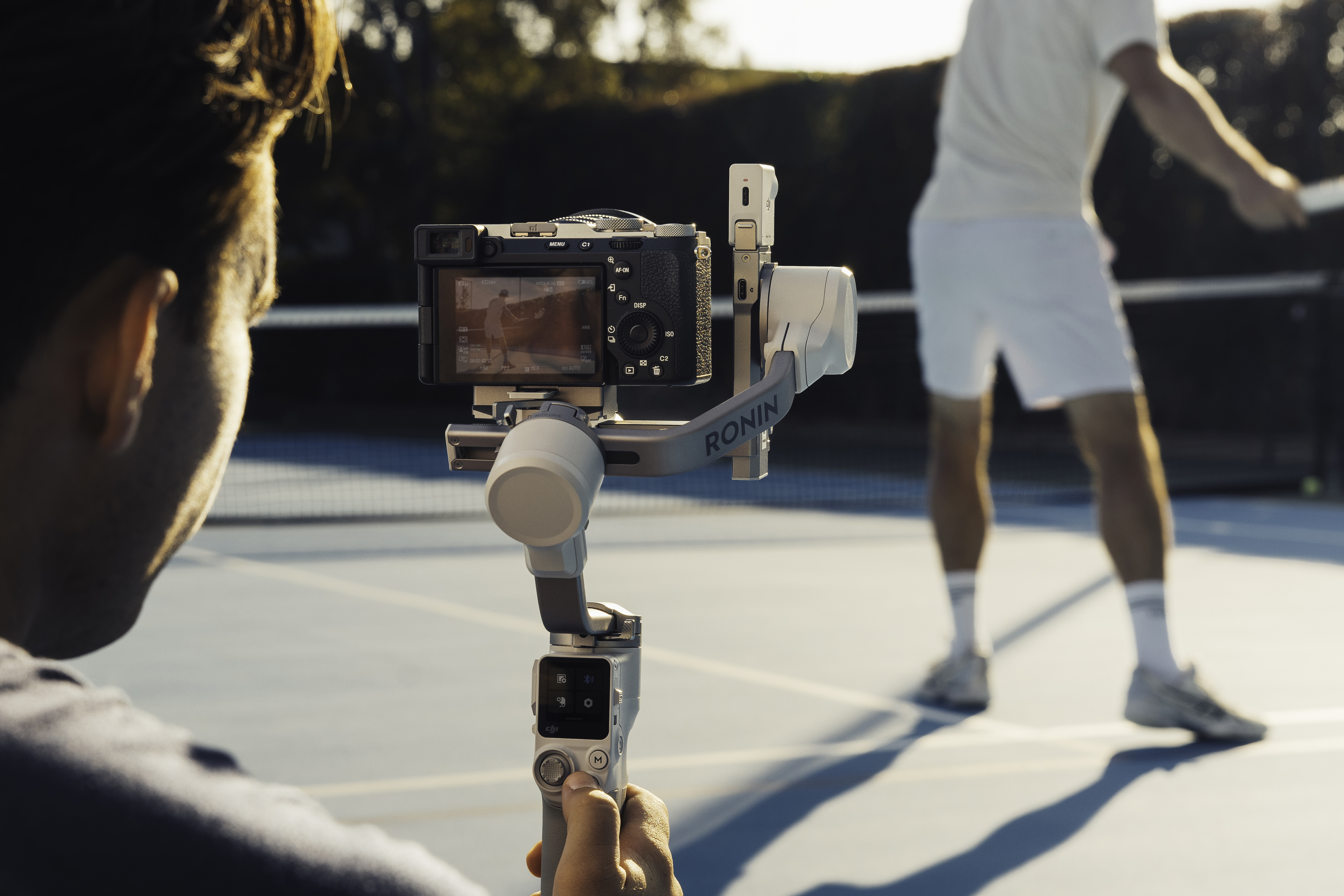 Une personne filme un match de tennis avec une caméra sur un cardan. Le match est en arrière-plan.